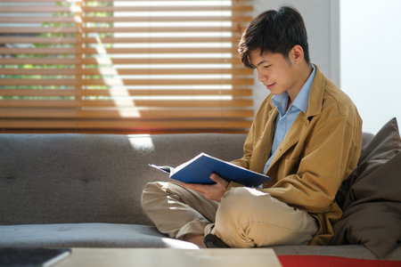 Calm asian man reading book in cozy bright living room with sunlight through blinds on windows .の写真素材