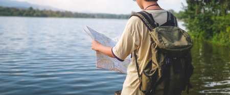 Young  male backpacker reading a map for direction while standing near river in sunny weather during...