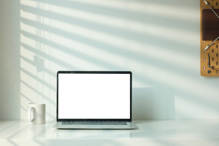 Front view of laptop computer and coffee cup on table with shadow from the jalousie on a white wall.の写真素材