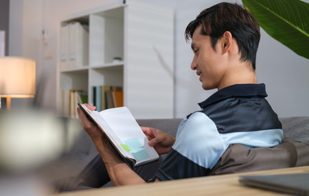 Happy millennial man sitting on couch in bright living room and reading book.