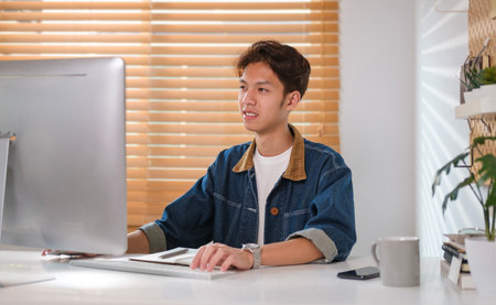 Smiling young asian man looking at computer screen, working online or browsing internet website.の写真素材