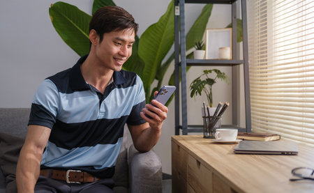 Portrait of smiling Asian male using smartphone sitting on the floor with laptop computer, reading text message, leaning on sofa, working at home officeの写真素材