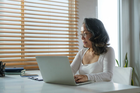 Beautiful mature woman in casual clothes sitting at table in home office and working on laptop computer.の写真素材