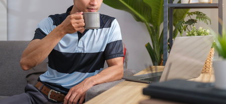 Cropped shot of asian man drinking hot coffee and reading email on laptop at home.の写真素材