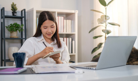 Smiling female entrepreneur taking notes, making business plans on notebook and using laptop at workplace.の写真素材
