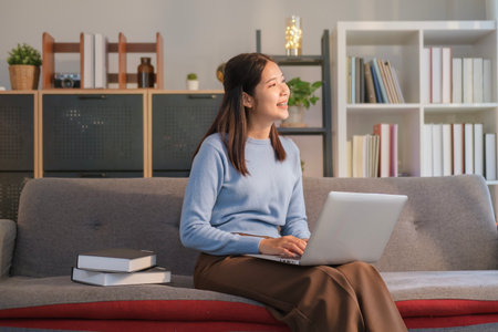 Happy young woman sitting on couch at home with laptop and looking through window.の写真素材