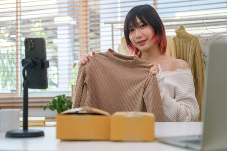 Pretty Asian female fashion vlogger presenting and showing clothes in front of smart phone, selling online live streaming.の写真素材