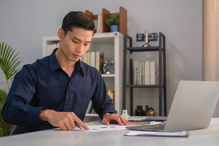 Concentrated asian male financial analyst working with financial report and using laptop at office.の写真素材