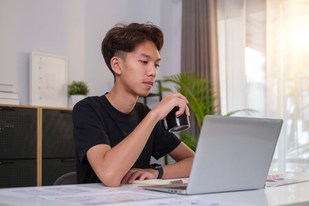 Image of young asian man drinking hot coffee and reading email or working online on laptop computer.の写真素材