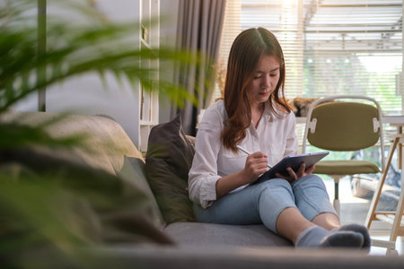 Full length of young woman in casual clothes sitting on couch and surfing internet on digital tablet.の写真素材