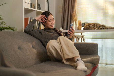 Happy Asian woman enjoys online chatting, shopping online on mobile phone while sitting on couch at home.の写真素材