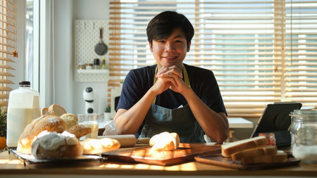 Portrait of handsome asian man sitting at kitchen table with freshly baked bread.の写真素材
