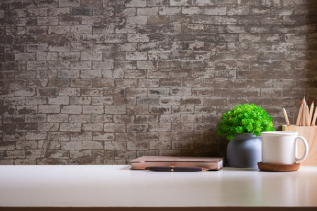 Stylish workspace with cup of coffee, book, houseplant and pencil holder on white table against brick wall.の写真素材