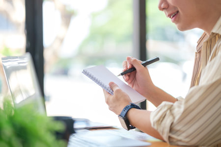 Smiling Asian man working on new project, looking at laptop and writing important notes.の写真素材