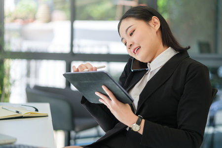 Smiling young businesswoman sitting in front of laptop at office desk and using digital tablet.の写真素材