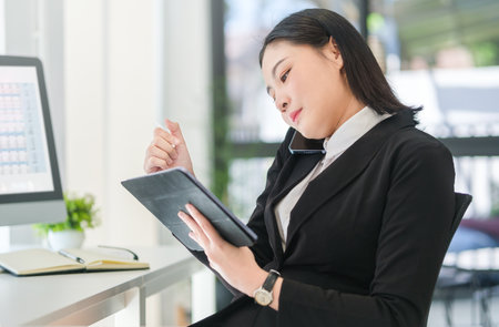 Busy young female employee having phone conversation and checking information on digital tablet.の写真素材