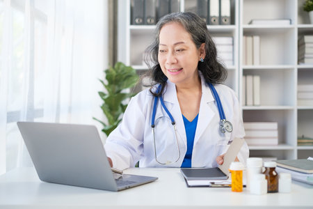 Shot of a female doctor wearing white coat writing filling medical form and using laptop at workplace.の写真素材