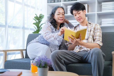 Happy middle age mother spending time with young adult son, reading book on couch at home.の写真素材