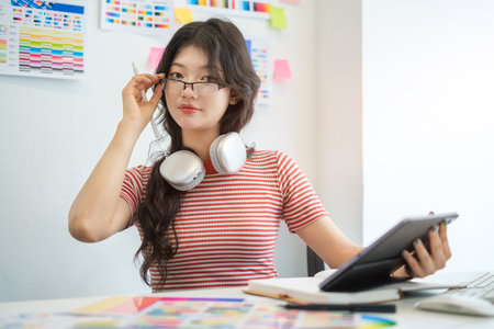 Confident young female designer working on digital tablet and choosing colorful paper charts at desk.の写真素材