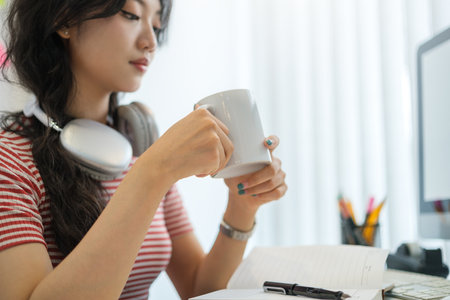Satisfied asian female freelancer sitting at workplace and drinking coffee.
