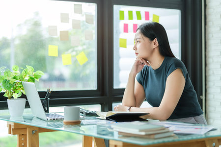 Pensive millennial business lady 
sitting at office desk and looking away thinking, contemplating the future.の写真素材