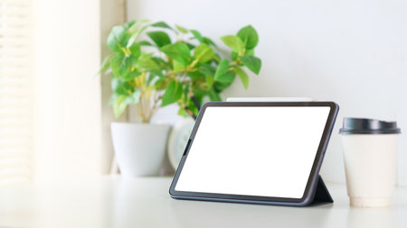 Digital tablet with blank display, paper cup of coffee and potted plant on white table. Modern workspace.の写真素材