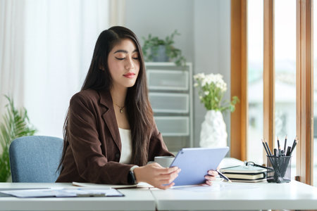 Beautiful millennial female entrepreneur sitting at workstation and reading email from client on digital tablet.の写真素材