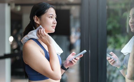 Smiling fitness woman wiping sweat with towel and using smartphone in gym.の写真素材