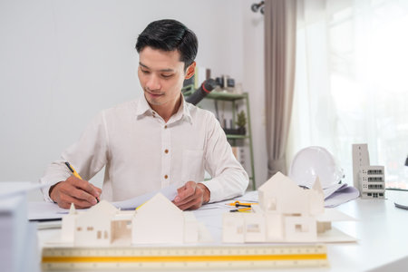 Handsome male architect working with blueprints, examining model of construction project at desk.の写真素材