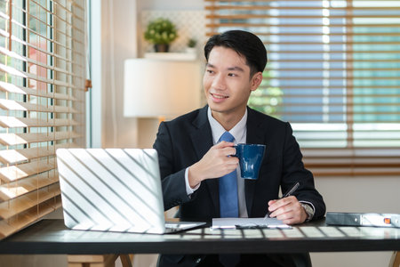 Relaxed asian man holding coffee and looking away while sitting in modern office.の写真素材