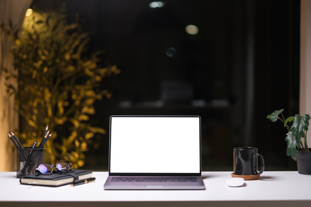 Laptop with blank screen coffee cup and potted plant on white table in modern dark office at night.の写真素材