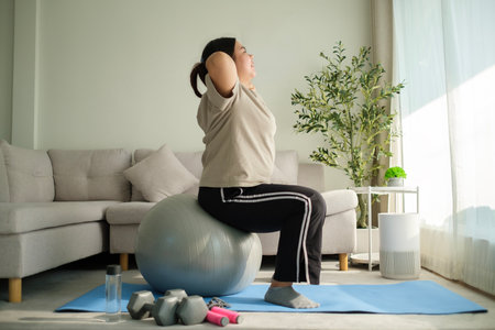 Full length of young overweight woman exercising with fitness ball on mat at home. Healthy lifestyle concept.の写真素材
