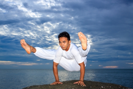Portrait of Asian young man doing yoga exercise on stone with cloudy sky and ocean, Thailandの写真素材
