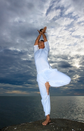 Portrait of Asian young man doing yoga exercise on stone with cloudy sky and ocean, Thailandの写真素材