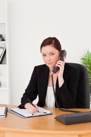 Young gorgeous red-haired woman in suit writing on a notepad and phoning while sitting in an officeの写真素材