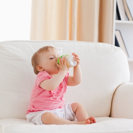 Lovely blond baby bottle-feeding while sitting on a sofa in the living roomの写真素材