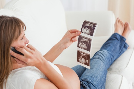 Attractive pregnant woman on the phone and looking at an ultrasound scan while lying on a sofa in her apartmentの写真素材