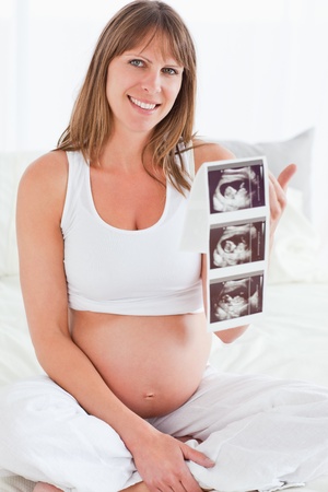 Beautiful pregnant female showing an ultrasound scan while sitting on a bed in her apartmentの写真素材