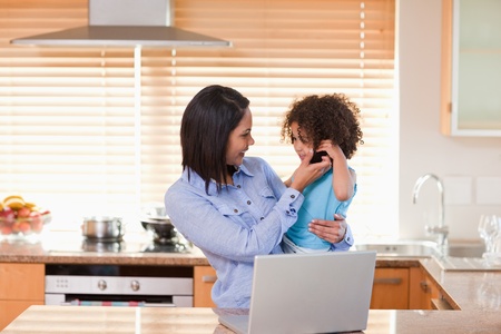 Young mother and daughter using laptop and cellphone in the kitchen togetherの写真素材