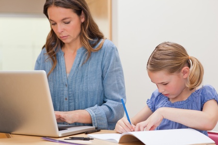 Girl doing her homework while her mother is using laptop in a kitchenの写真素材