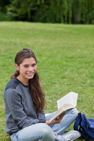Young smiling girl sitting cross-legged on the grass while holding a book in a parkの写真素材