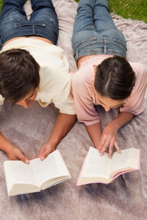 Man and a woman reading books while lying prone on a grey blanket in the grassの写真素材