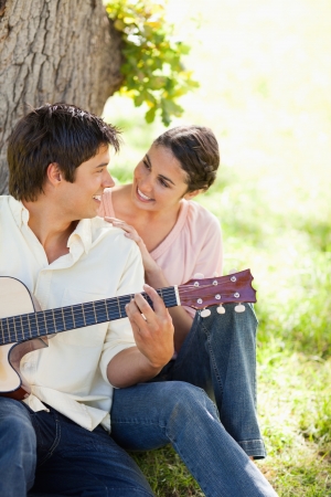 Smiling woman looking eye to eye with her friend who is holding a guitar as they both sit against a treeの写真素材