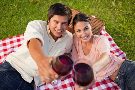 Man and a woman smiling happily as they touch their raised glasses of red wine during a picnic with focus on the glasses of wineの写真素材