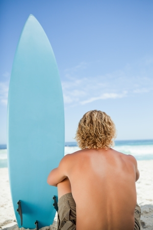Blonde man sitting down with his arms around his legs next to his surfboardの写真素材