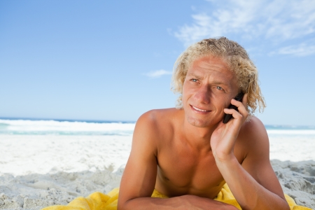 Blonde man lying on his beach towel while talking on his mobile phoneの写真素材