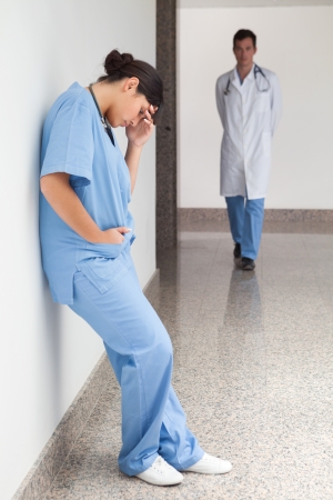 Sad urse leans against wall in hospital corridor with doctor approachingの写真素材