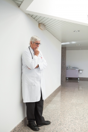 Thinking doctor leaning against wall with had holding chin in hospital corridorの写真素材