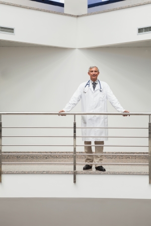 Doctor standing at the railing of the hospital corridor smilingの写真素材