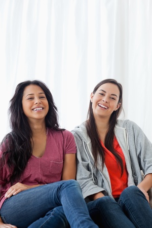 A pair of laughing young ladies sit on the couch as they look into the cameraの写真素材